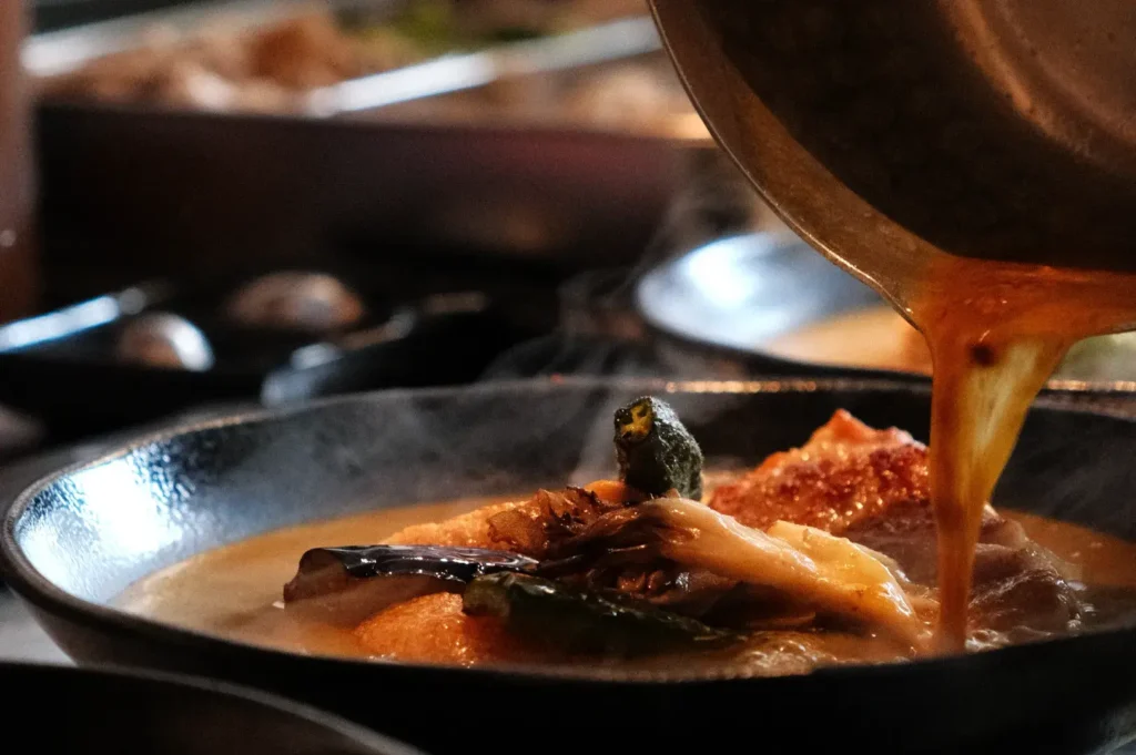 A close-up of a steaming dish being served, featuring vibrant vegetables and broth being poured from a pot into a bowl.