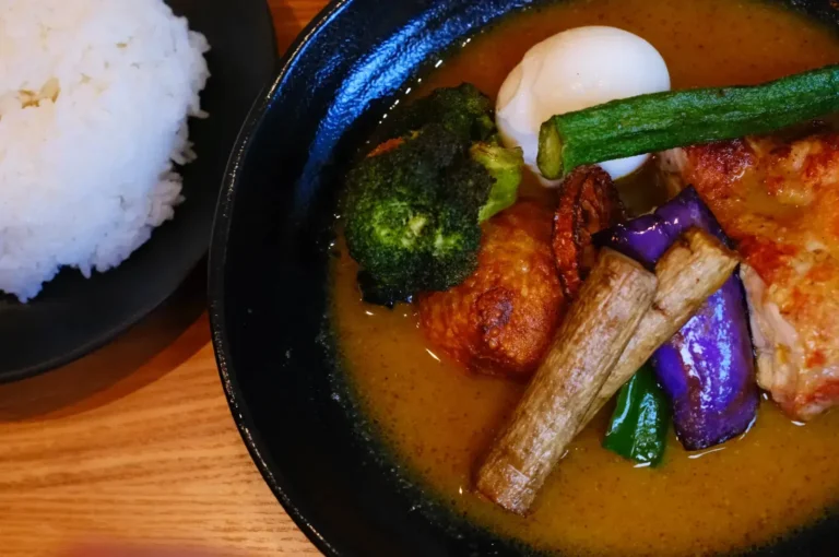 A bowl of curry with various vegetables, including broccoli, okra, and eggplant, alongside a soft-boiled egg and fried chicken, served with a side of white rice.