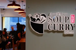 Restaurant interior with warm pendant lights, diners at tables, and a wall sign reading “Soup Curry by Ki-Kosui” beside a bowl logo.