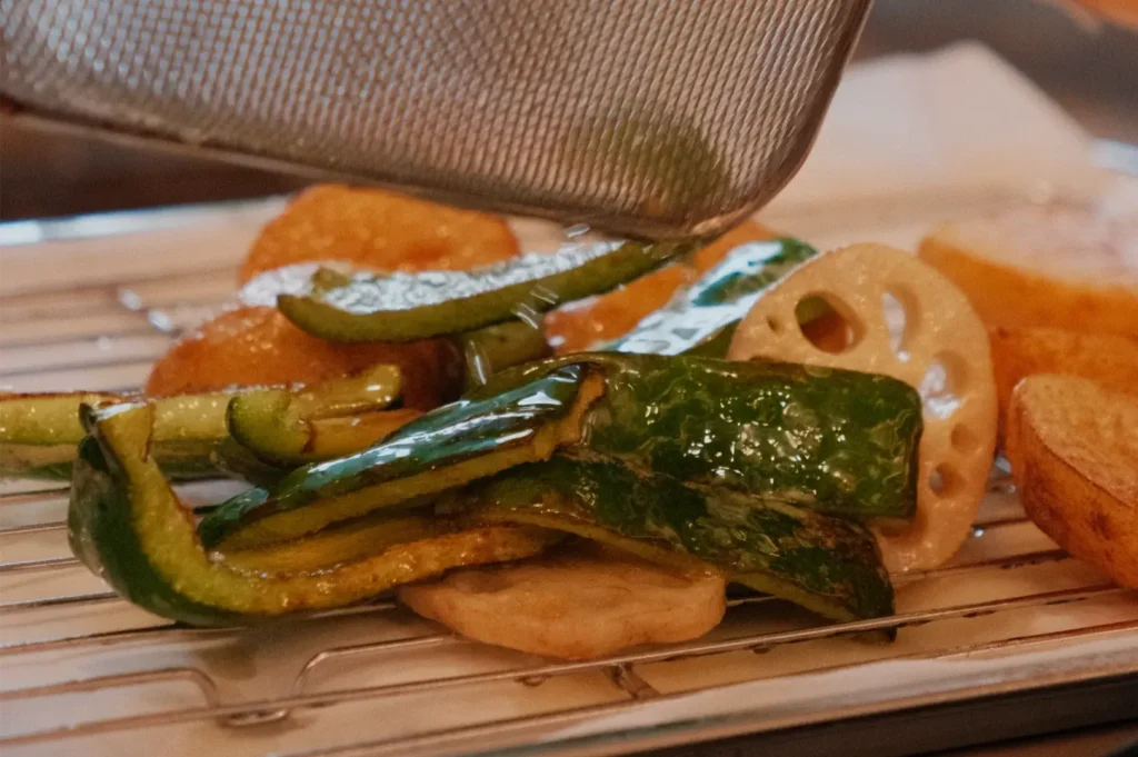 A mesh strainer lifts over golden fried vegetables, revealing crisp green pepper, zucchini, and lotus root on a wooden draining rack; warm, appetizing tones.