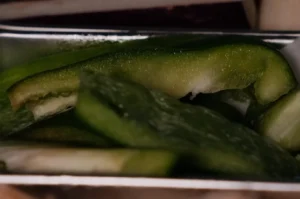 Close-up of sliced green pickles stacked in a metal tray, their glossy skins and pale interiors visible in dim light, creating a quiet, moody tone.