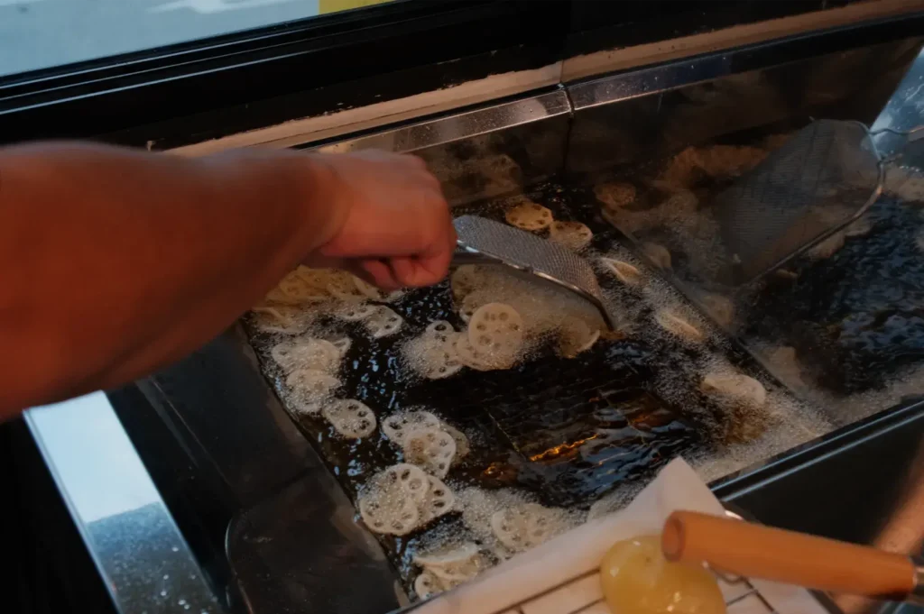 A hand uses a metal skimmer to turn round battered snacks in a bubbling commercial deep fryer, with sizzling oil and a busy kitchen feel.