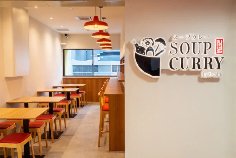 Brightly lit interior of a Japanese restaurant showing small wooden tables, counter seating, and a large backlit wall logo reading "Soup Curry By Ki-setsu."