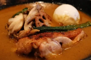 A rich orange curry surrounds browned chicken, mushrooms, lotus root, a green pepper, and a soft-boiled egg in a dark bowl, warm and hearty.