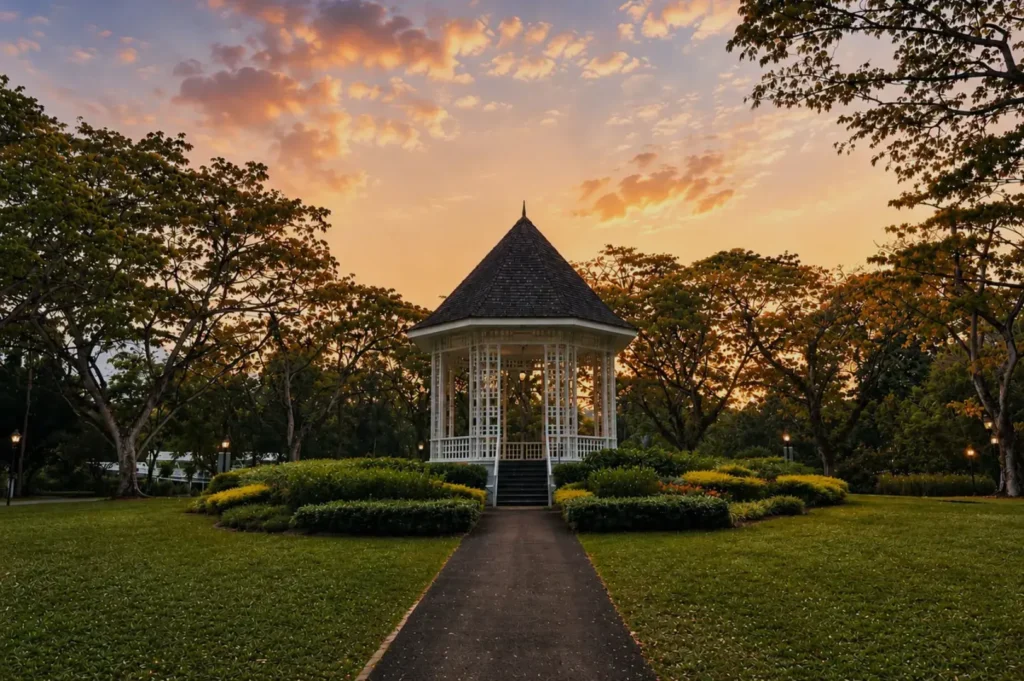 A white gazebo with a dark pointed roof stands at the end of a garden path, framed by trees and flower beds beneath a glowing sunset sky.