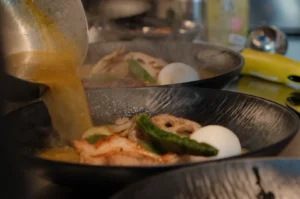 Hot broth pours from a ladle into a black bowl of ramen topped with shrimp, green vegetables, lotus root, and a soft-boiled egg; warm, inviting kitchen scene.