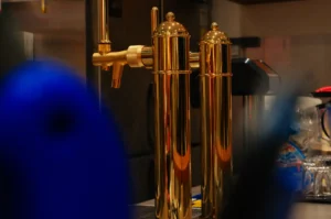Two polished brass beer taps gleam against a dim bar backdrop, framed by a blurred blue figure in the foreground, creating a warm, lively pub atmosphere.