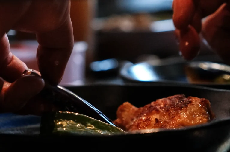 Hands use metal tongs to turn a golden-brown piece of food beside a green vegetable in a skillet, capturing a focused, intimate cooking moment.