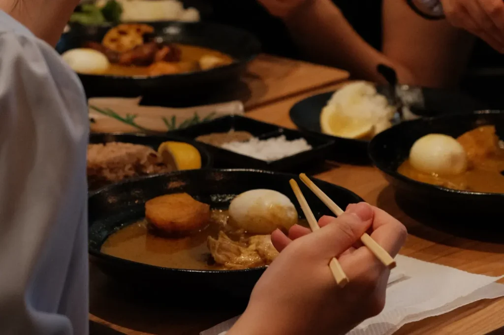 A diner holds chopsticks over a black pan of broth with dumplings and fish balls, surrounded by shared dishes at a warmly lit table.