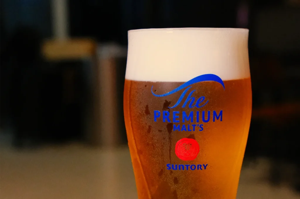 A chilled glass of amber Suntory The Premium Malt’s beer, topped with thick white foam and condensation, glows against a dim, warm bar.