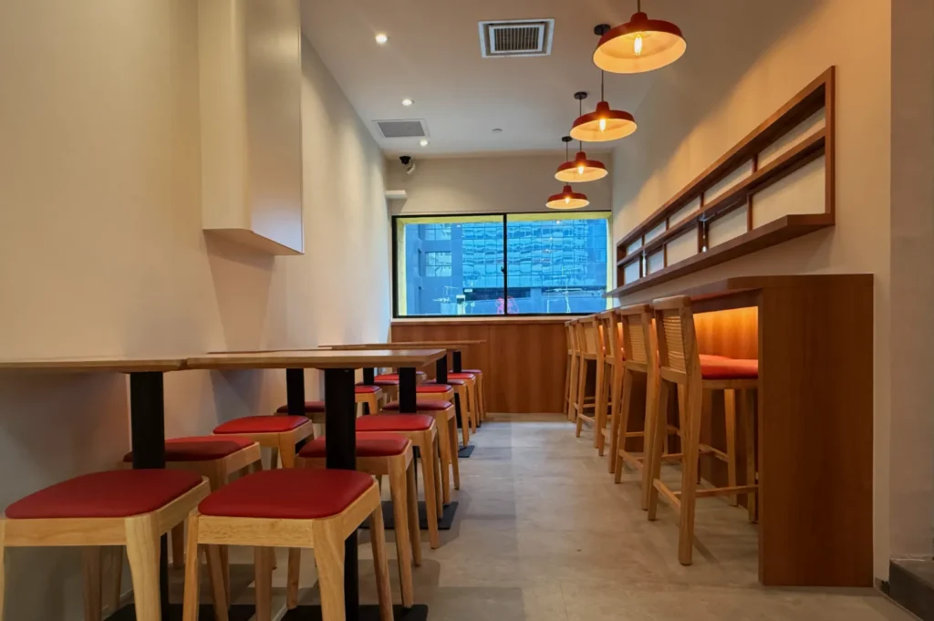 A narrow café with wooden tables and red-cushioned stools, a long bar, glowing pendant lights, and a city view through the rear window.
