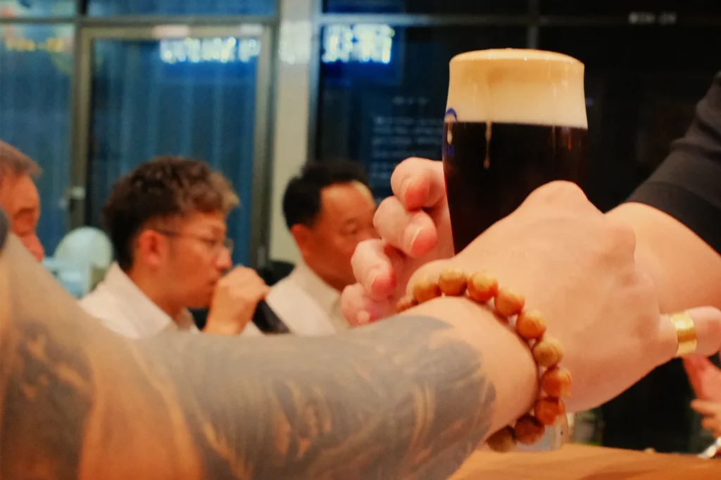 Tattooed hands and arms raise a dark stout beer with a creamy foam head in a toast, while diners chat in a warmly lit restaurant.