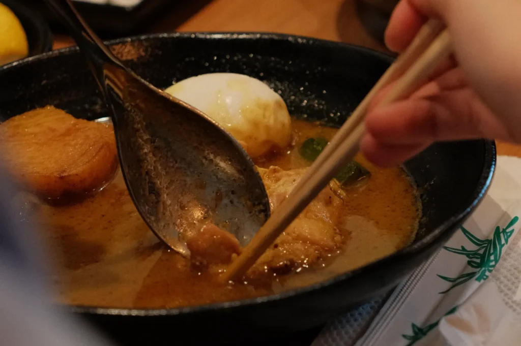 A hand uses chopsticks and a spoon to lift food from a black bowl of rich Japanese curry, served with a soft-boiled egg and vegetables.