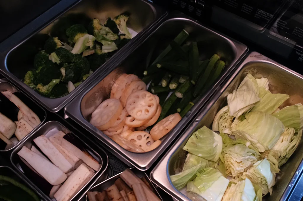 Stainless-steel buffet trays display broccoli, okra, lotus root slices, cabbage, and other vegetables, creating a fresh, abundant meal spread.