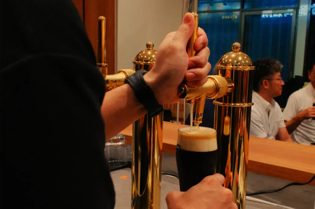 Tattooed hands and arms raise a dark stout beer with a creamy foam head in a toast, while diners chat in a warmly lit restaurant.