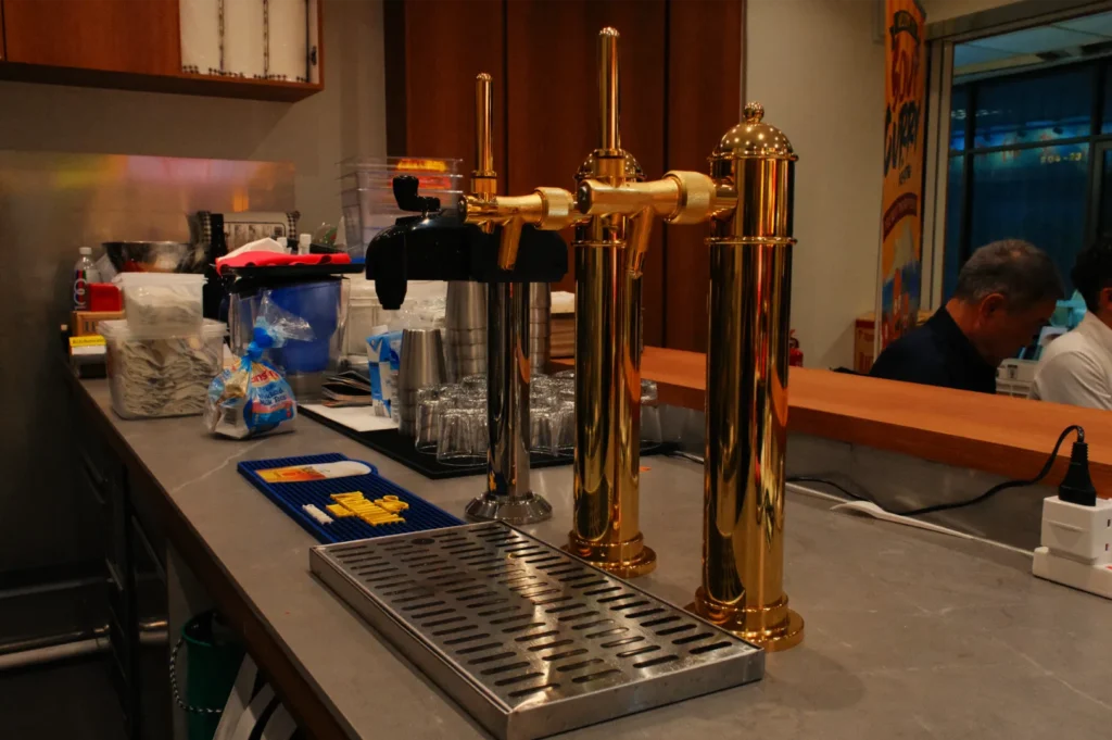 Polished brass beer taps stand on a bar counter beside a metal drip tray, with café supplies and staff visible in the warm-lit background.