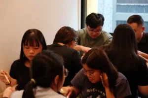 A small group of young adults gathers around a table, eating and talking over food in a bright indoor setting; the mood is casual and friendly.