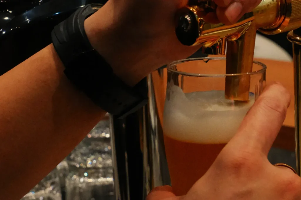 A bartender pours amber beer from a tap into a glass, building a foamy white head in a dimly lit bar; the close-up feels warm and lively.