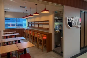 Warm, modern soup curry restaurant with wooden tables, red pendant lights, and a long bar lined with stools; a “SOUP CURRY” sign marks the entrance.