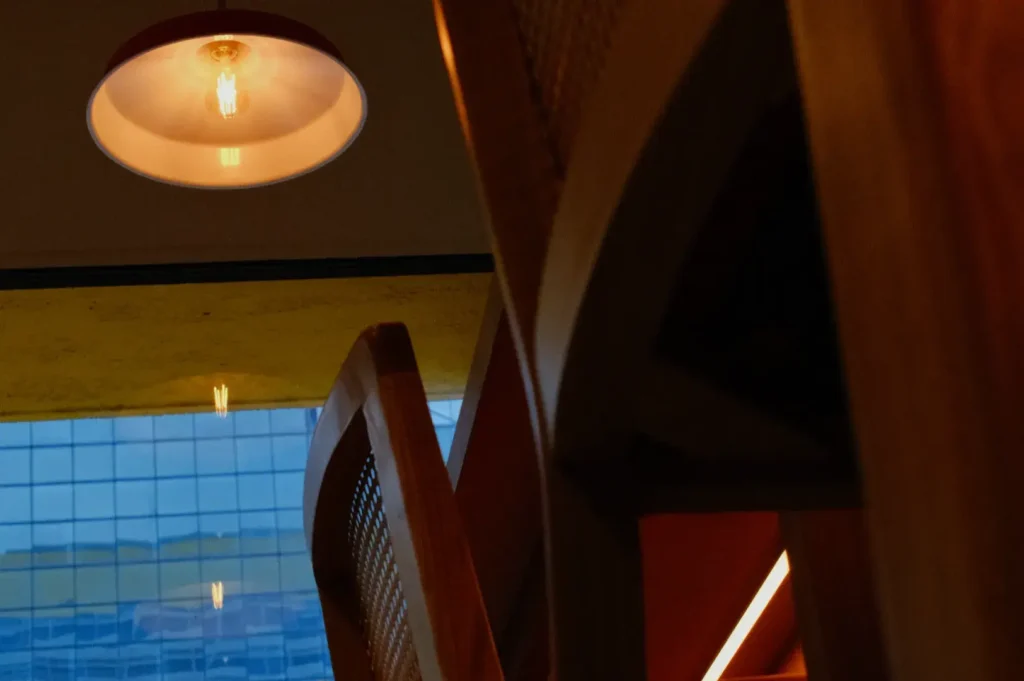 Warm ceiling light glows above a modern interior with sweeping wooden arches, a red accent panel, and blue glass windows, creating a moody architectural atmosphere.