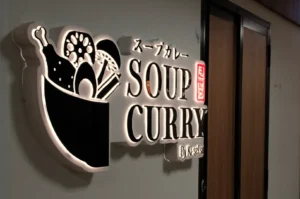 Illuminated sign for a Japanese soup curry restaurant, with a bowl-and-vegetable graphic and bold “SOUP CURRY” lettering beside a wooden doorway. Warm, inviting tone.