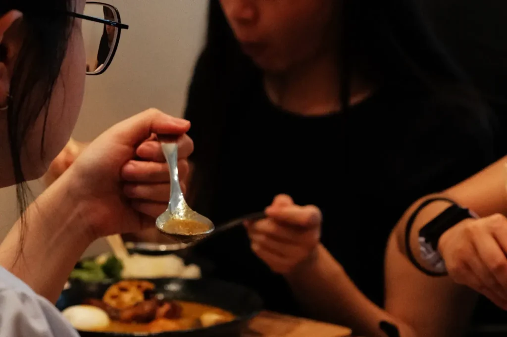 A person wearing glasses tastes food from a spoon while another diner gestures beside a meal; the warm, intimate restaurant setting feels relaxed and social.