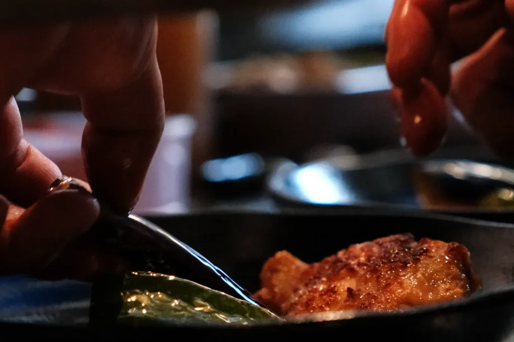 Hands season a browned piece of meat in a hot skillet, adding herbs or spices; warm, close-up lighting gives the cooking scene an intimate feel.