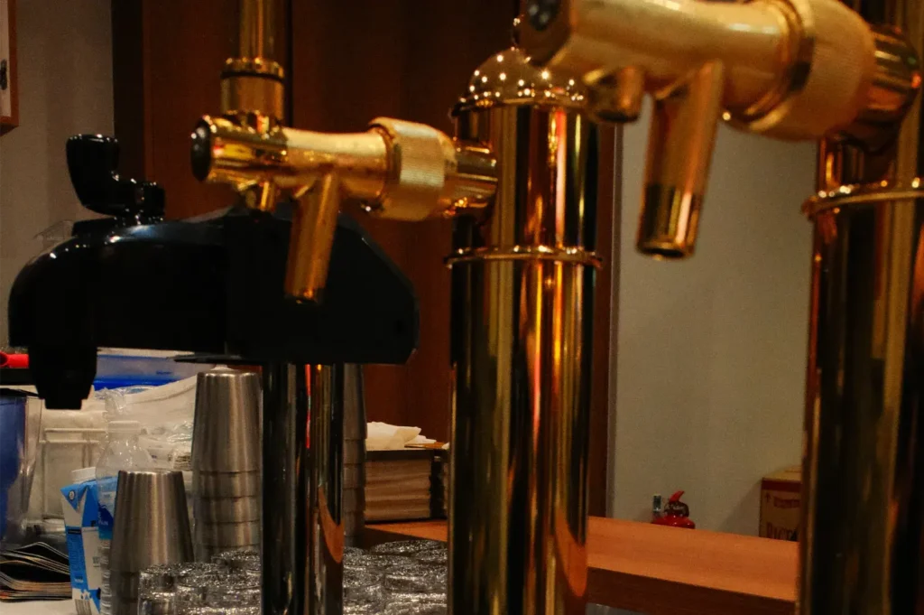 Polished brass beer taps dominate a warmly lit bar, with stacked glasses and brewing equipment behind them, creating a cozy, inviting pub atmosphere.