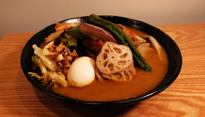 A bowl of soup curry with various toppings, including a soft-boiled egg, lotus root, asparagus, and vegetables in a rich broth.