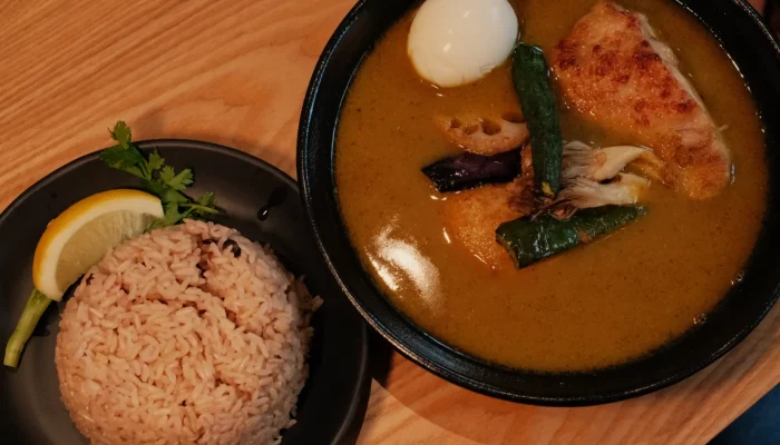 A bowl of curry with chicken and vegetables, accompanied by a serving of rice on a plate, garnished with a lemon wedge and cilantro.