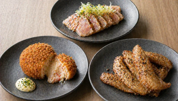 Gourmet Japanese appetizers featuring a creamy crab croquette with tartar sauce, seared tuna tataki, and peppery grilled chicken wings.