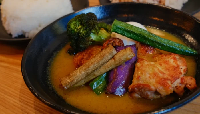 A bowl of soup curry with chicken, broccoli, green beans, eggplant, and okra, served with rice in the background.