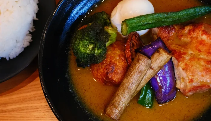A bowl of curry with pieces of chicken, a boiled egg, various vegetables including broccoli and eggplant, and a side of rice.
