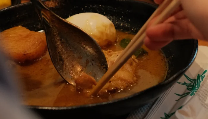 A close-up of a bowl of ramen being served with chopsticks, featuring slices of meat, a soft-boiled egg, and broth.